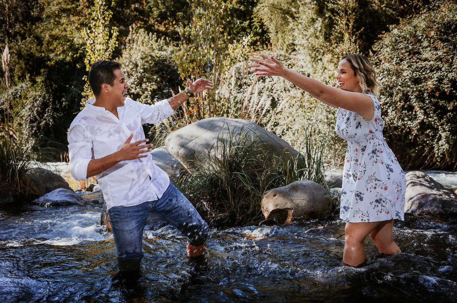 A man and woman playfully splash in a rocky river, reaching toward each other, wearing casual summer clothes outdoors.