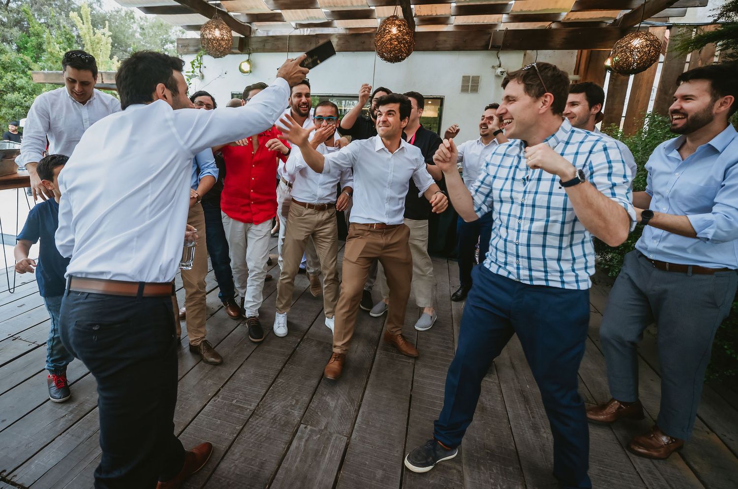 Group of men dancing and celebrating at an outdoor party on a wooden deck.