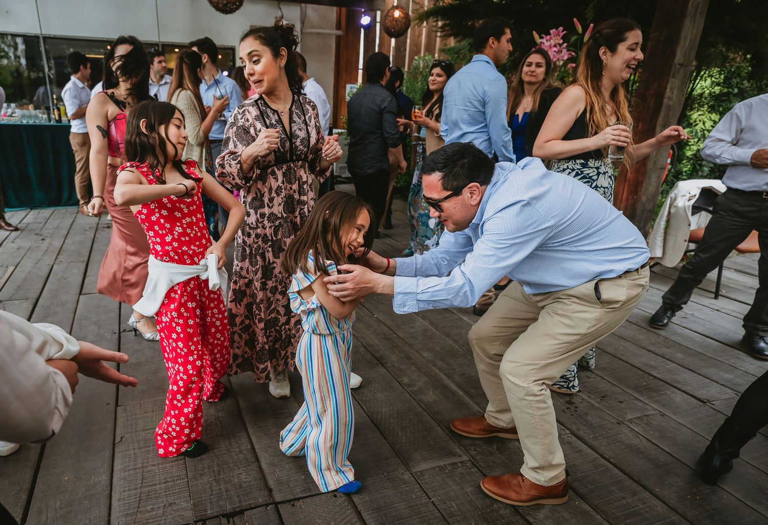 Adults and children dancing joyfully on a wooden deck at an outdoor celebration or wedding reception.