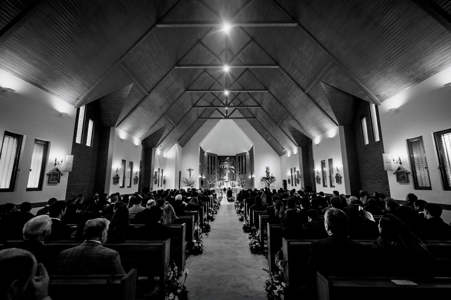 Black and white photo of a wedding ceremony inside a church with guests seated in pews and altar lit up.