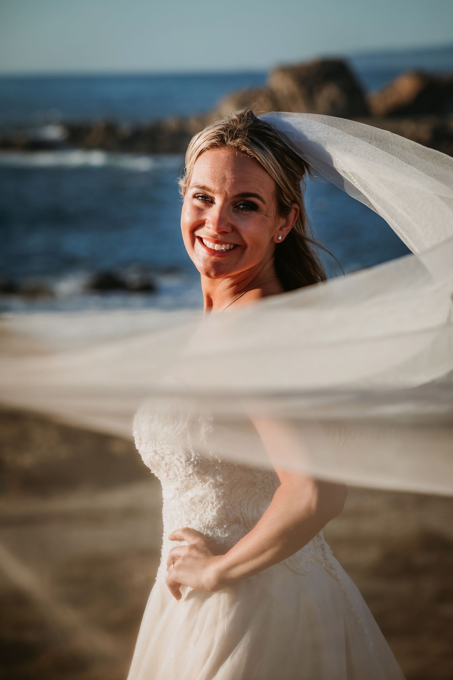 Smiling bride with veil blowing across her face on a sunny rocky beach.