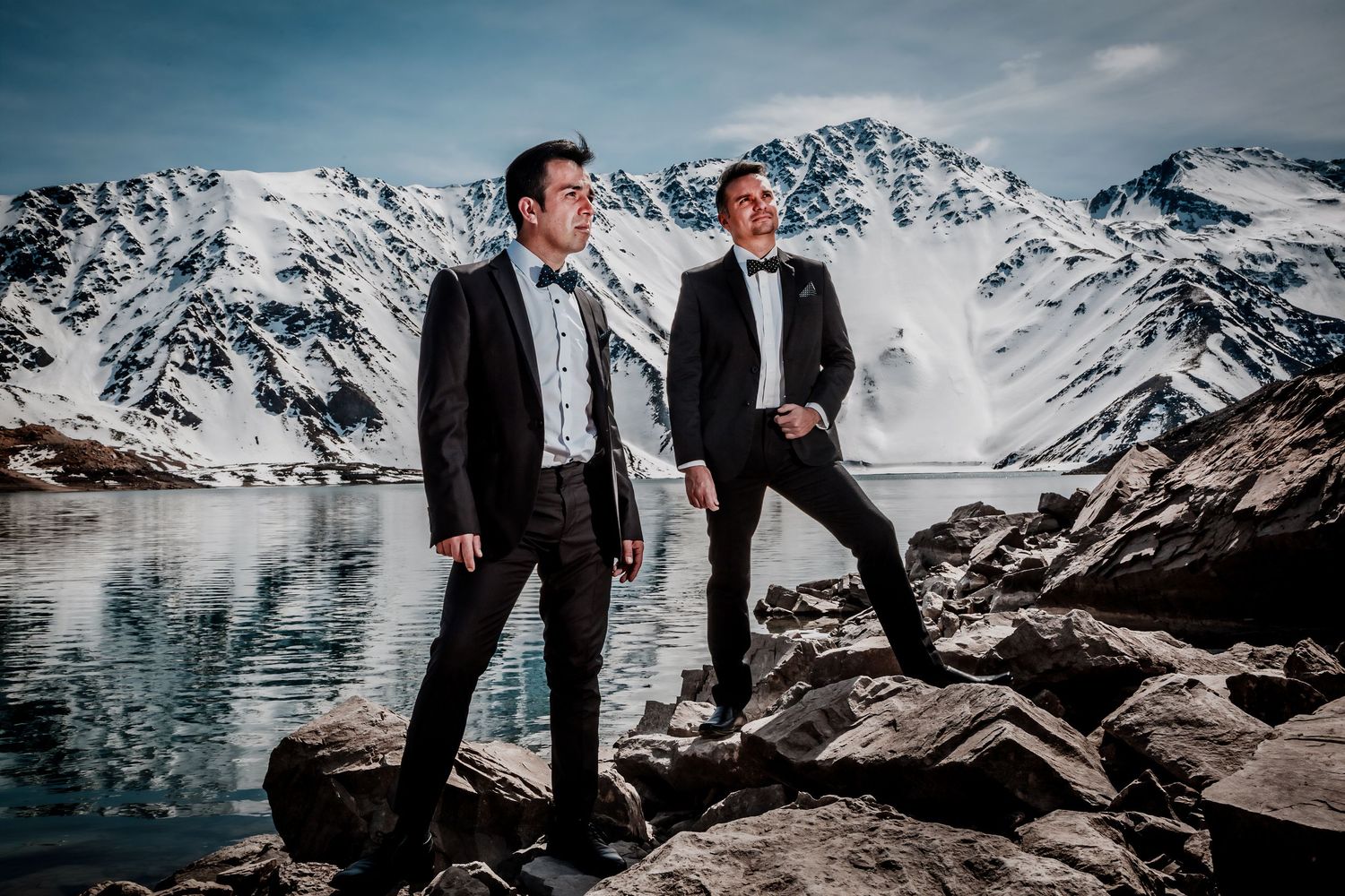 Two men in formal tuxedos stand on rocky shore with snow-covered mountain lake backdrop.
