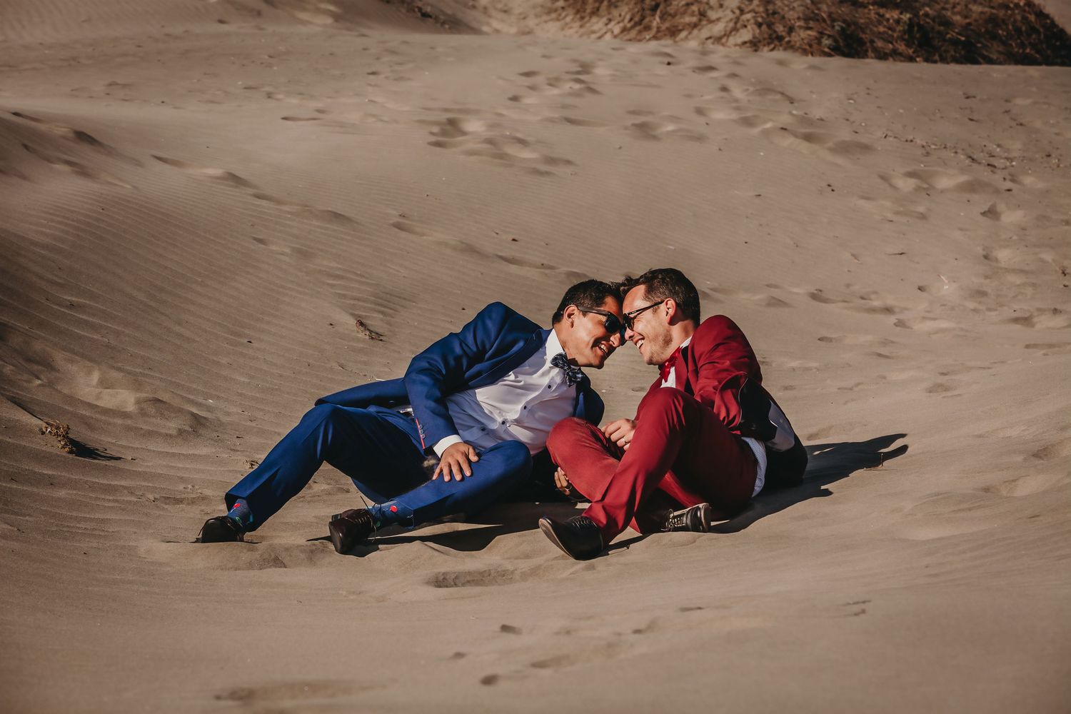 Two grooms in colorful suits sitting intimately together on sun-drenched sand dunes.