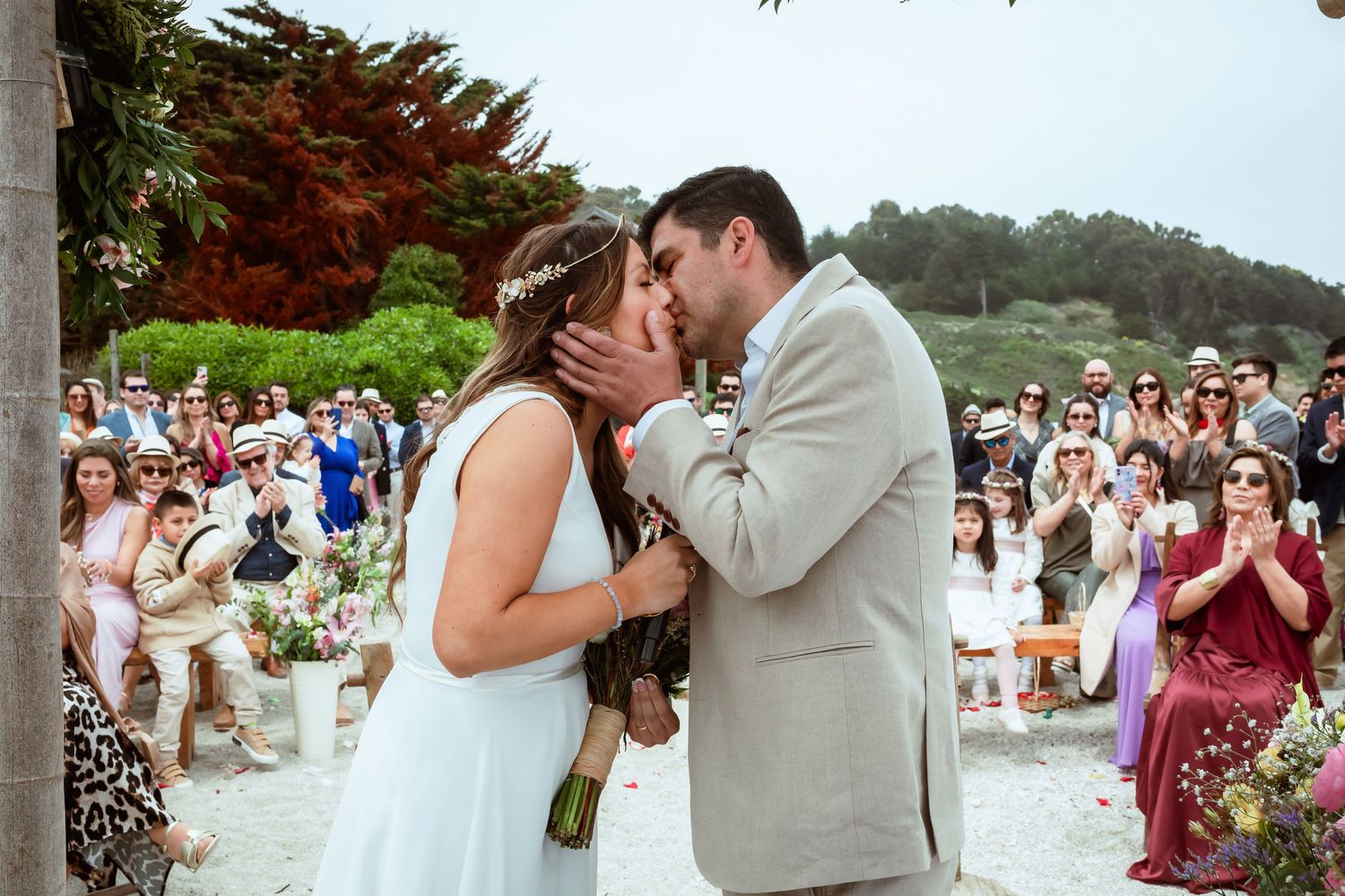 Bride and groom share first kiss as newlyweds while guests applaud at outdoor beach wedding.