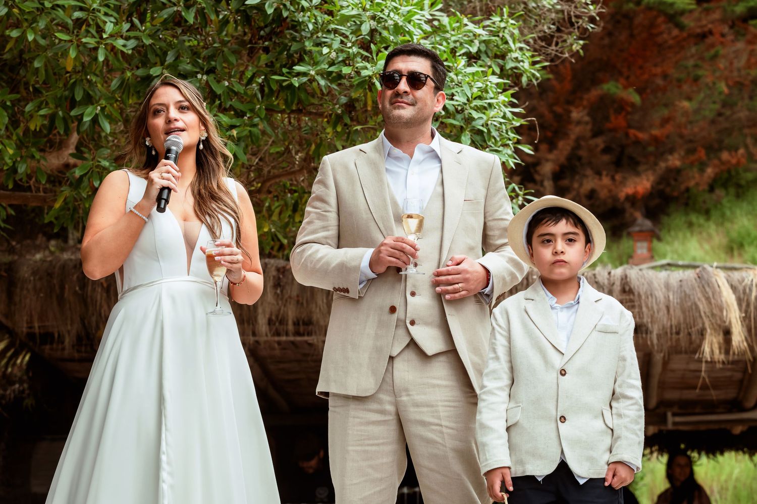 Bride speaks into microphone beside groom and young boy in white suit at outdoor wedding reception.