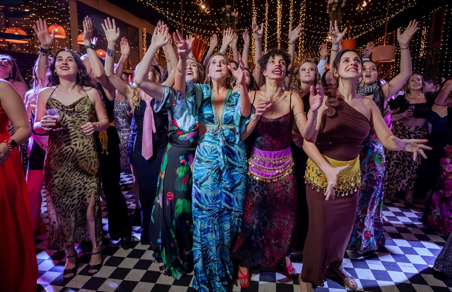 Wedding guests in elegant dresses dancing with arms raised on checkered floor at festive reception.