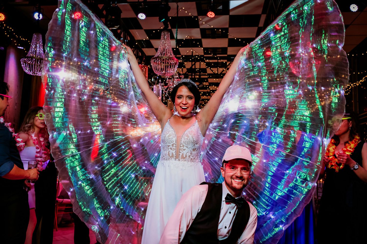 Smiling bride with illuminated rainbow LED wings posed with groom at wedding celebration.