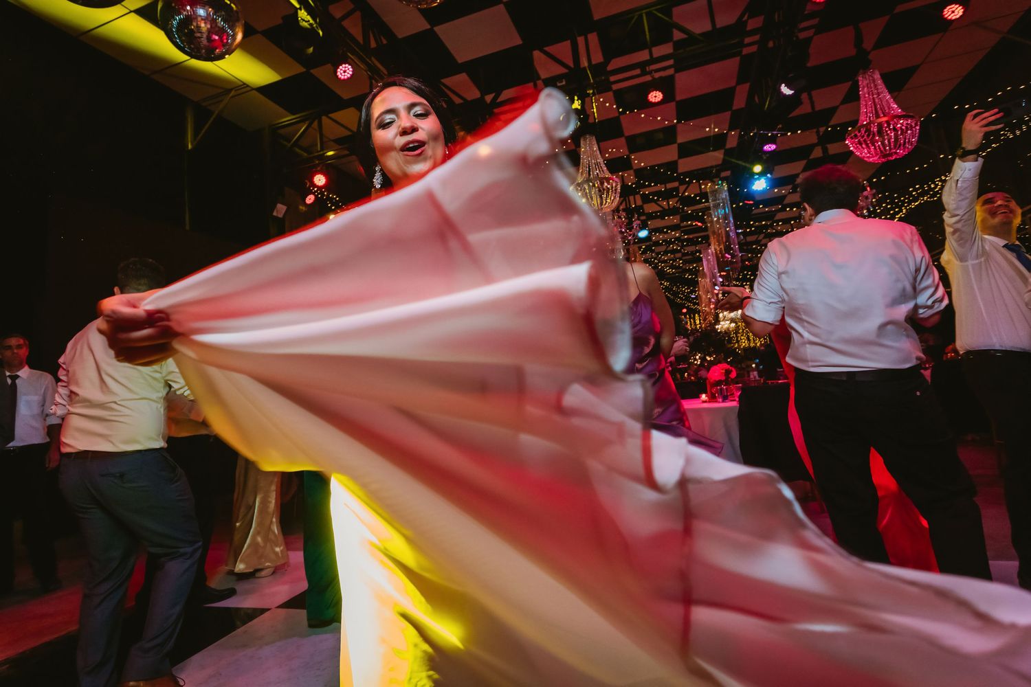 Guest twirls flowing pink fabric on crowded wedding reception dance floor under warm lights.