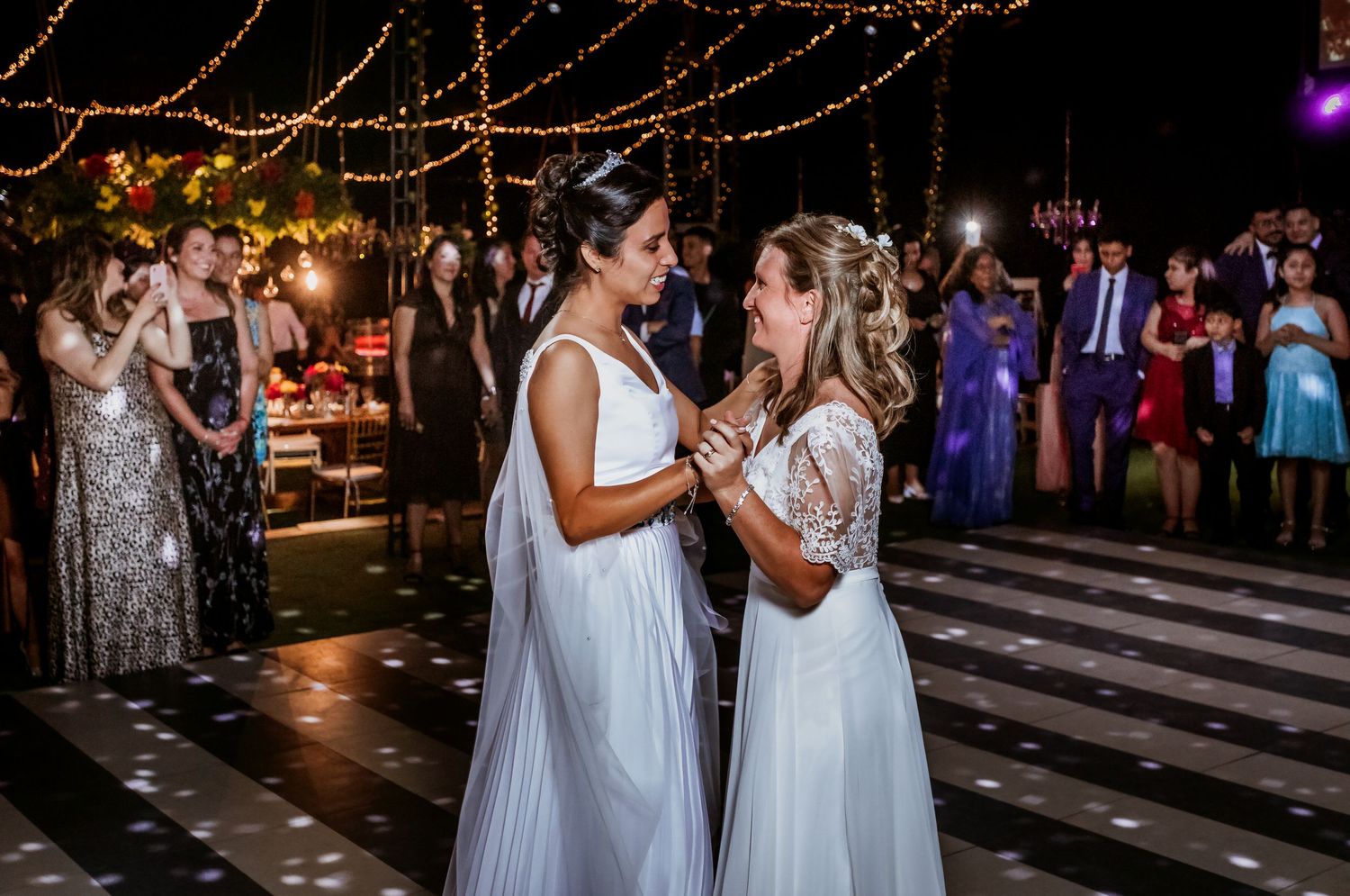 Two brides in white gowns share an intimate dance moment at outdoor evening wedding reception.