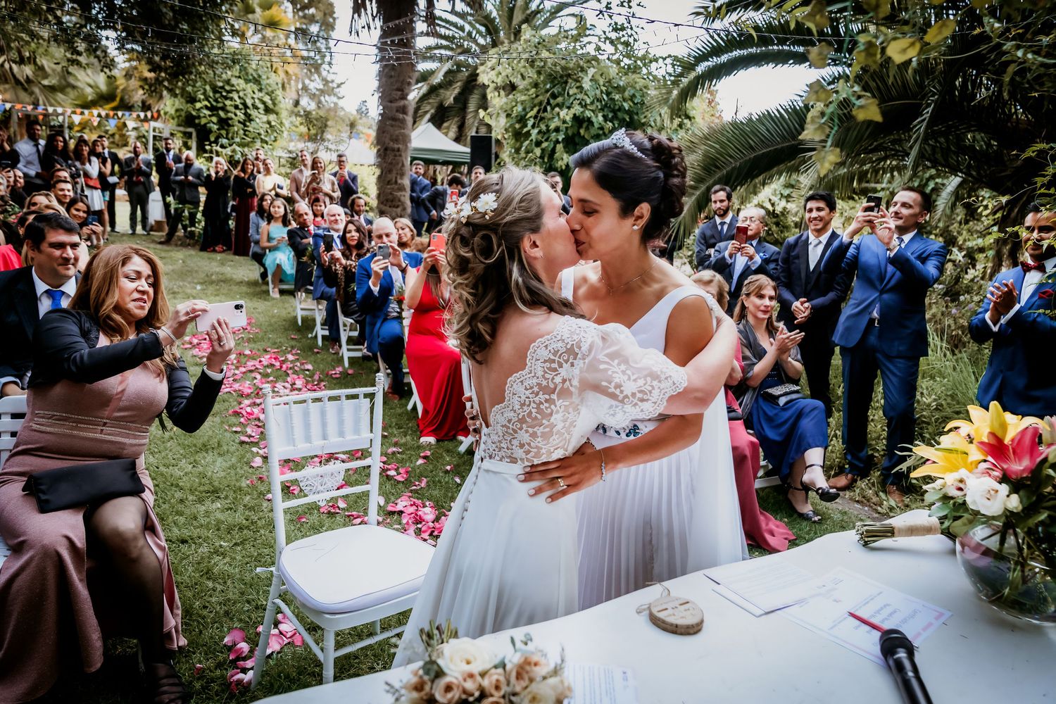 Two brides share their first kiss as a married couple as guests cheer and applaud.