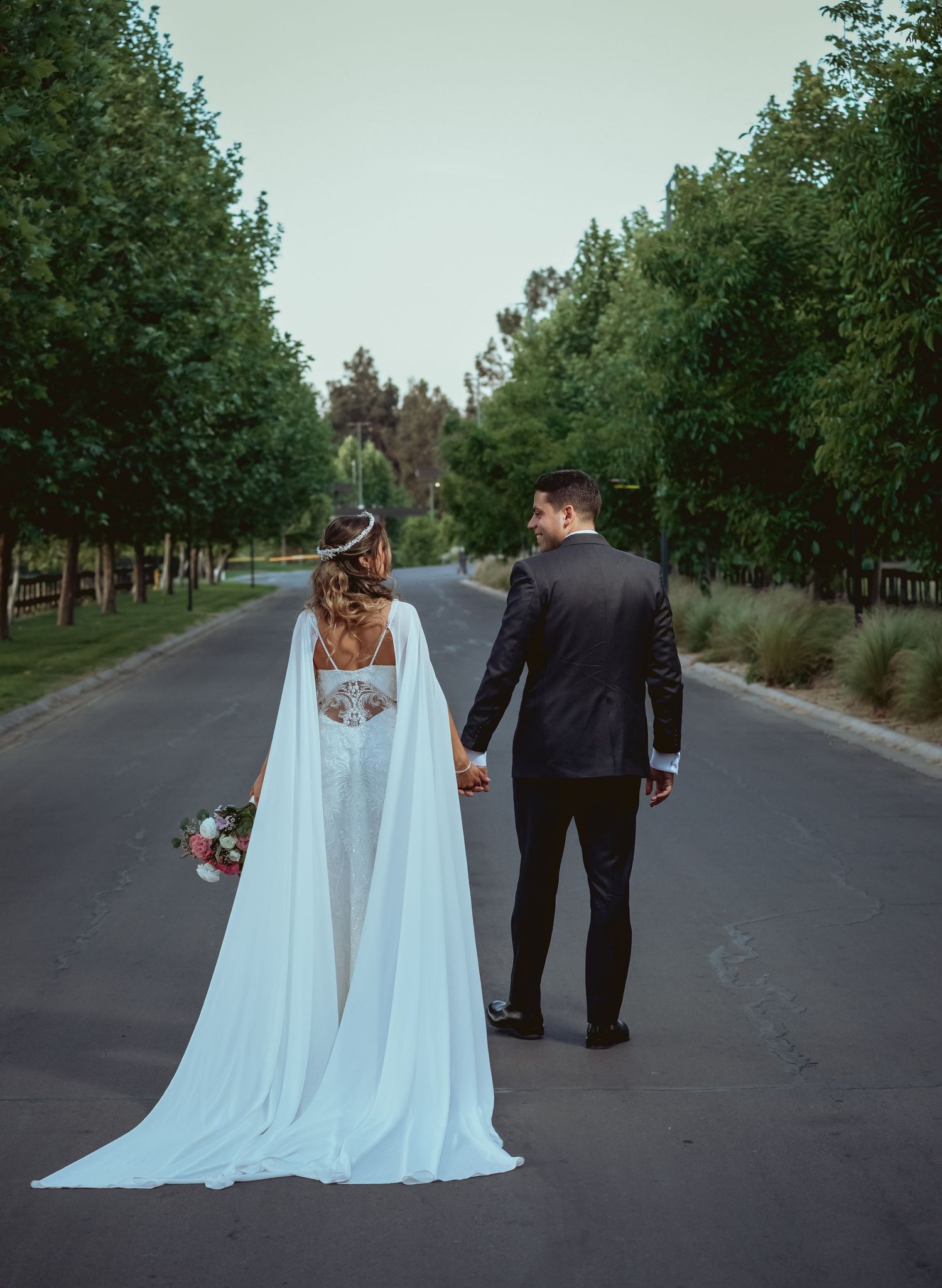 Bride in white cape gown and groom walking hand in hand down a tree-lined road at sunset.