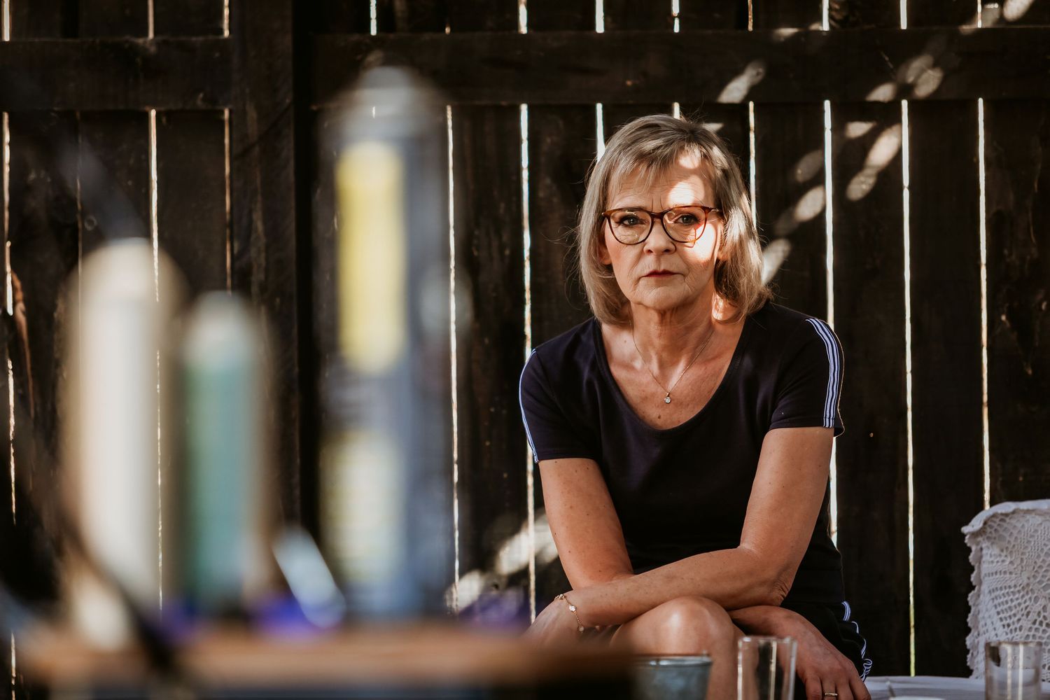 Mature woman with glasses sitting pensively in a rustic wooden barn setting.