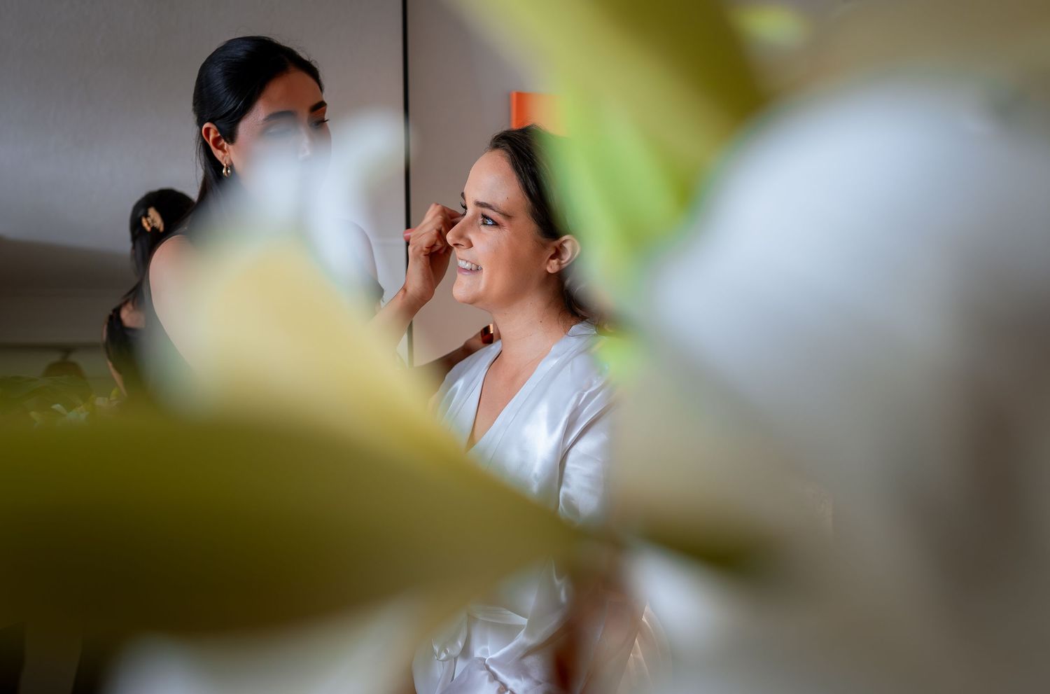 Bride in white robe getting makeup applied, photographed through soft-focus white flowers.