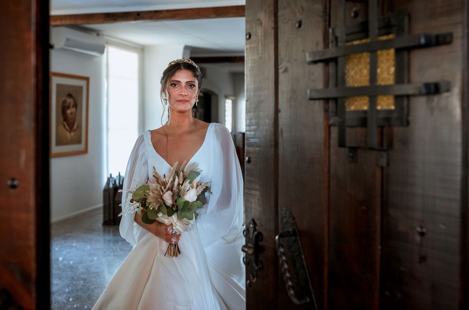 Elegant bride in long-sleeve white gown holding a boho bouquet standing in a rustic doorway.
