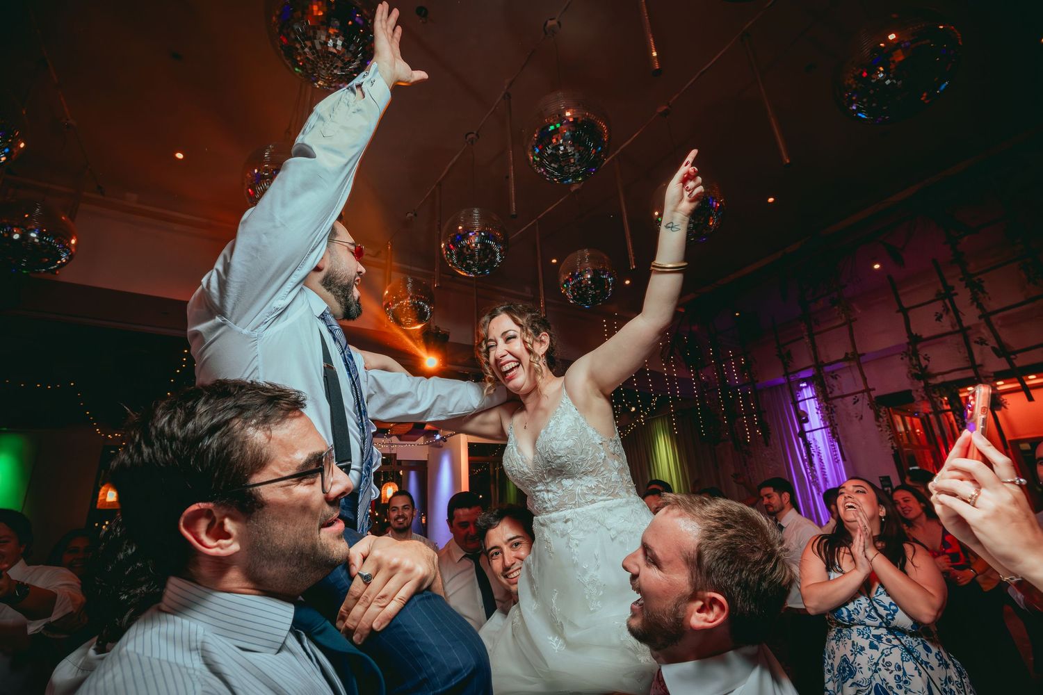 Excited bride lifted by wedding guests while celebrating on a festive dance floor.