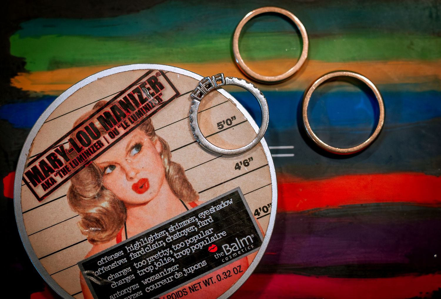 Wedding rings and bracelet displayed on colorful makeup compact by theBalm cosmetics brand.