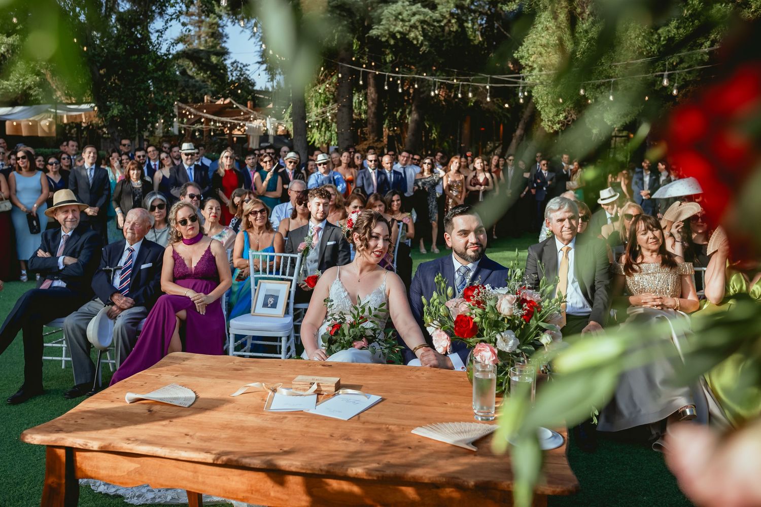 Bride and groom sit at rustic wooden table during outdoor garden wedding with guests seated behind.