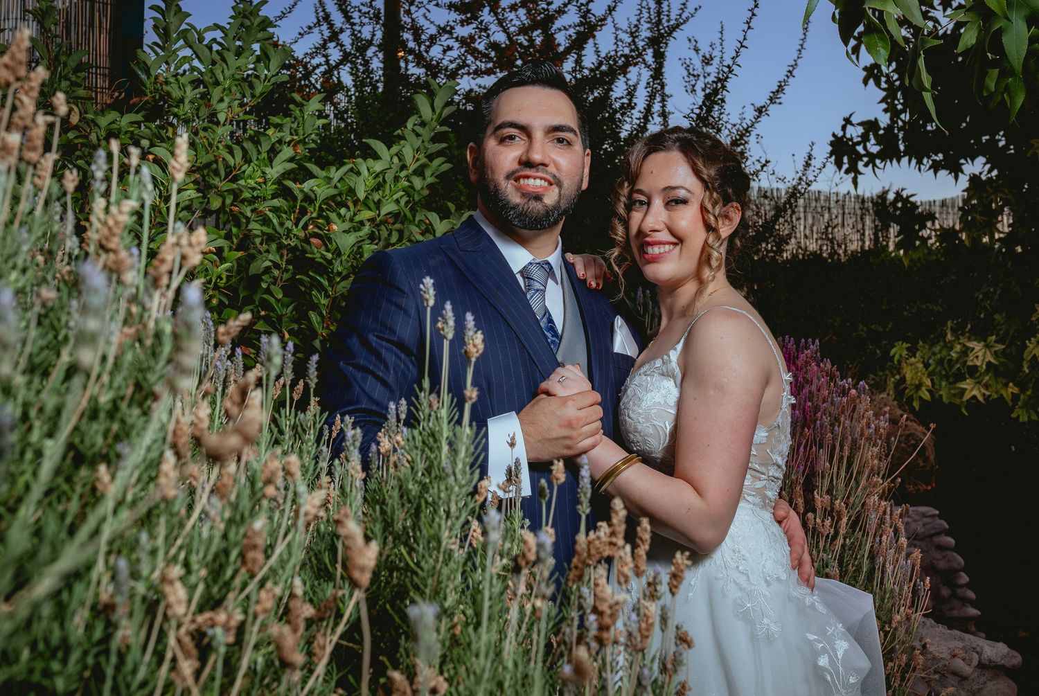 Smiling bride and groom pose together among lavender plants in a sunlit garden.