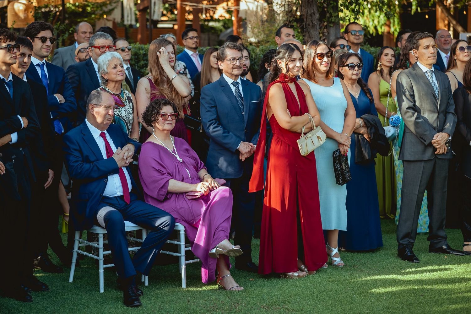 Elegantly dressed wedding guests stand and watch outdoor ceremony in formal attire on sunny day.