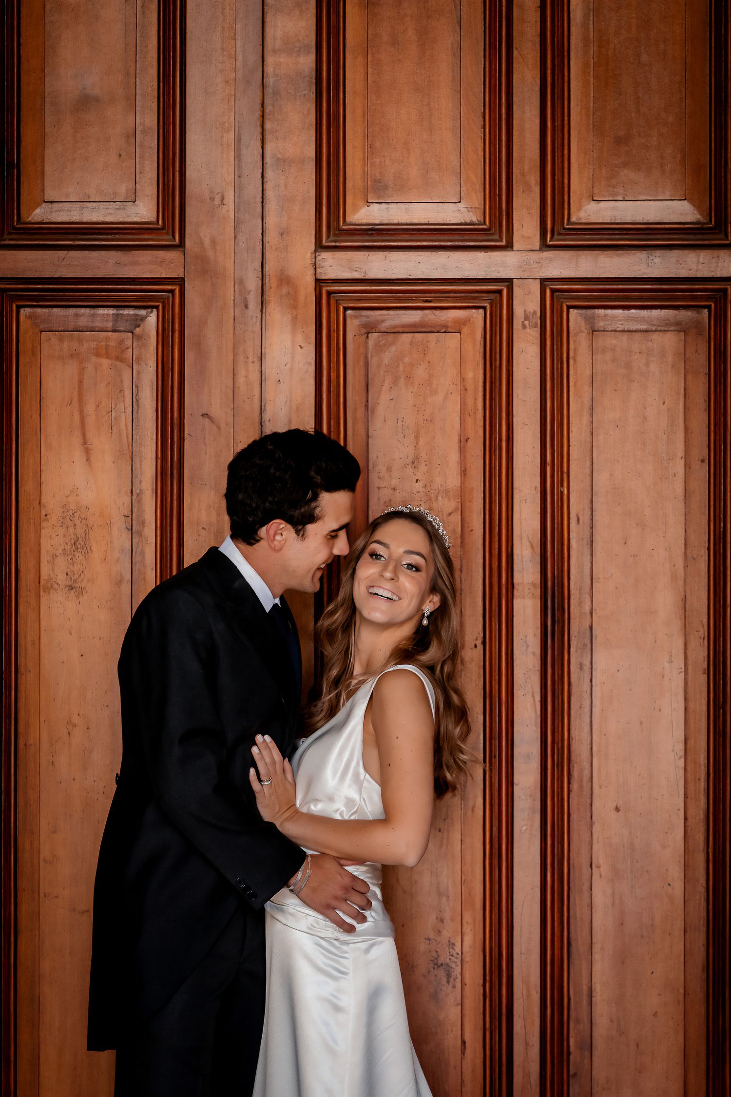 Groom whispering to smiling bride as they embrace against a grand wooden door backdrop.