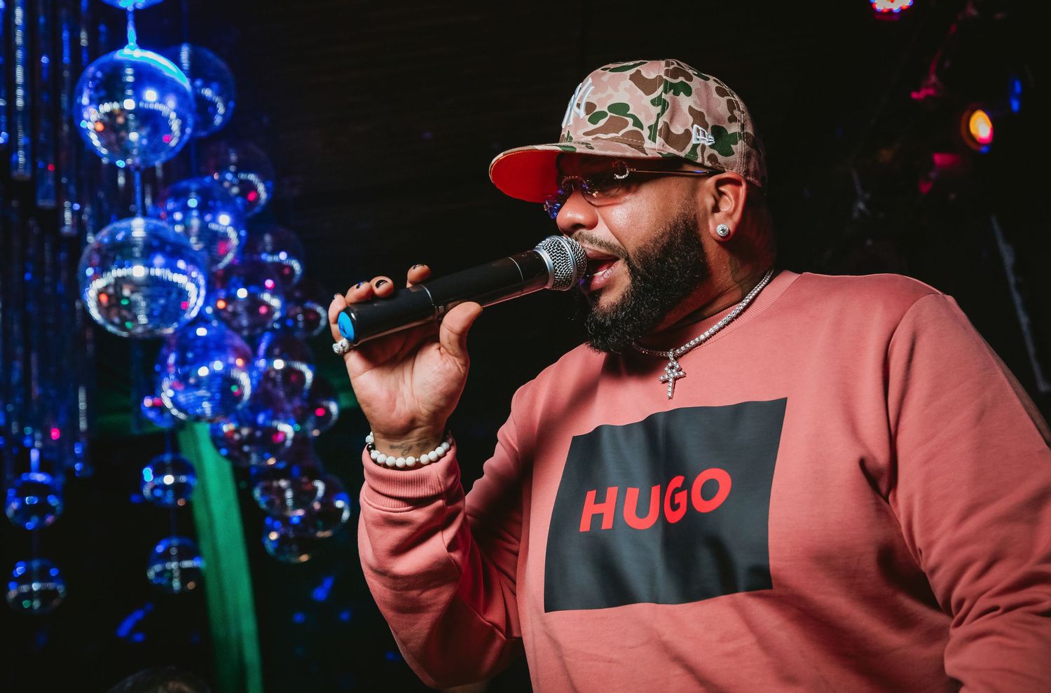 A male performer wearing a Hugo Boss pink sweatshirt and camo cap sings into a microphone at a glamorous event.