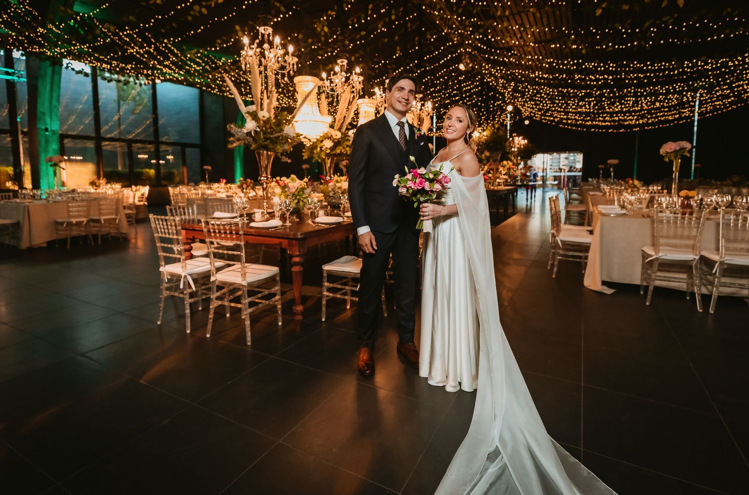 Elegant bride and groom pose in beautifully decorated wedding reception venue with fairy lights.