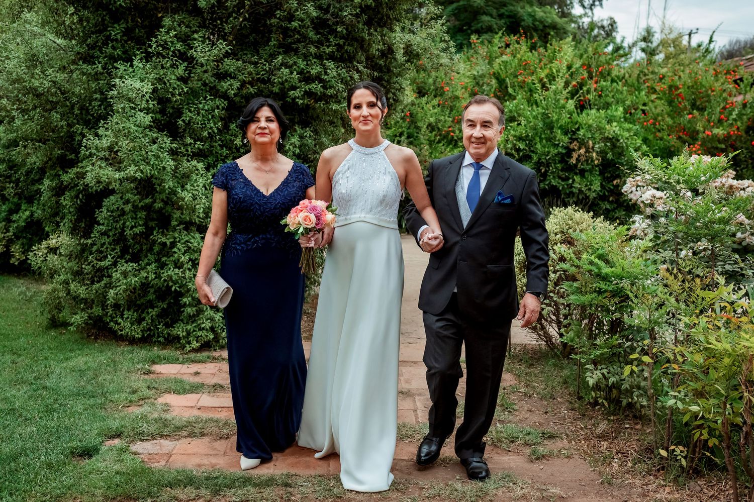 Bride in light blue gown walking with parents through garden path before wedding ceremony.