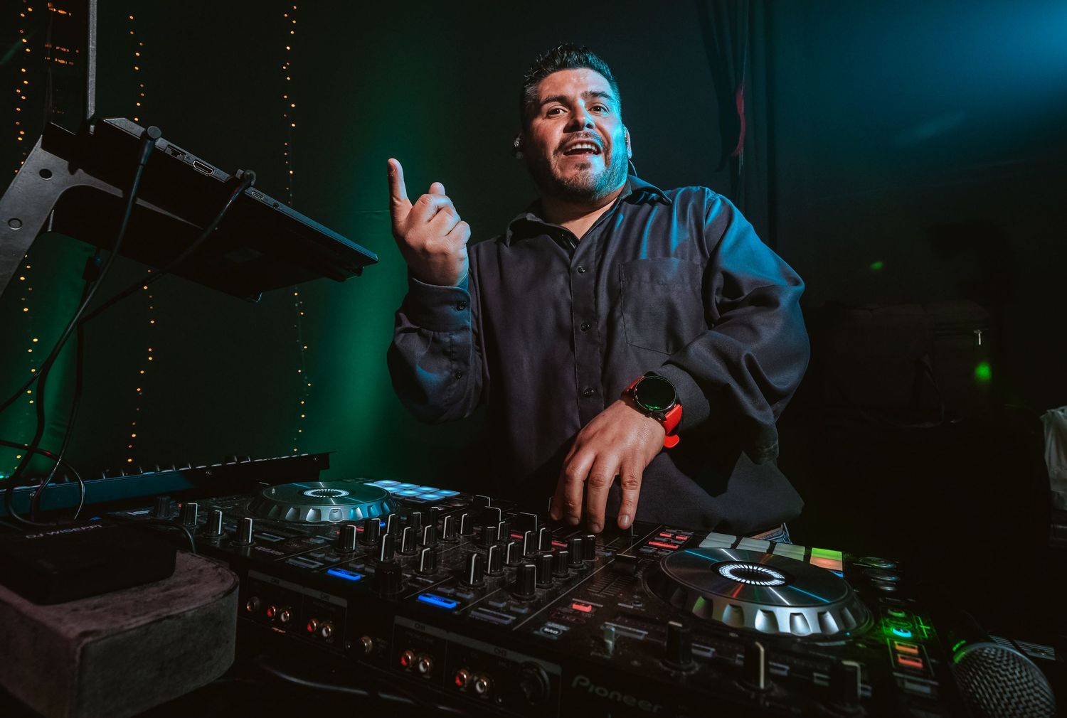 DJ performing at a nightclub, smiling and gesturing behind a mixer console under green stage lighting.