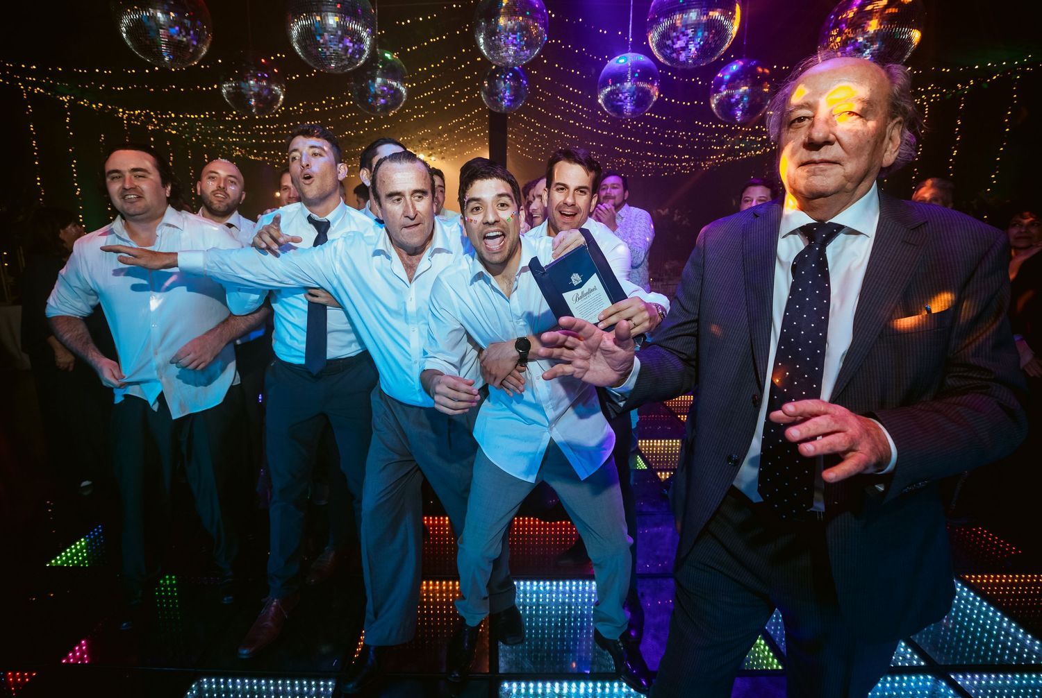 Group of men in white shirts dancing at a nightclub with disco balls and colorful lights.