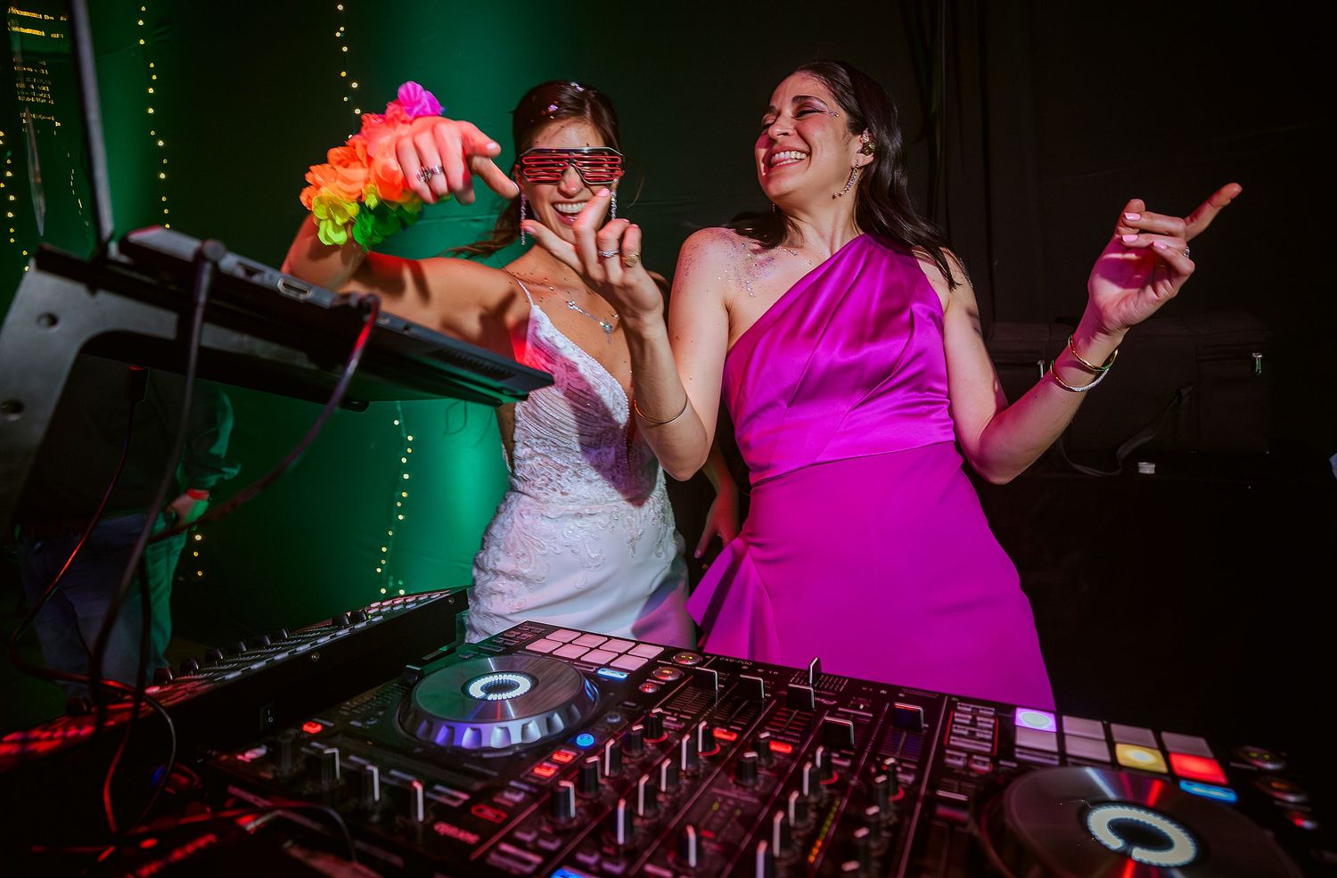 Two women laugh and dance behind a DJ booth at a wedding reception, one in white bridal gown, one in pink dress.