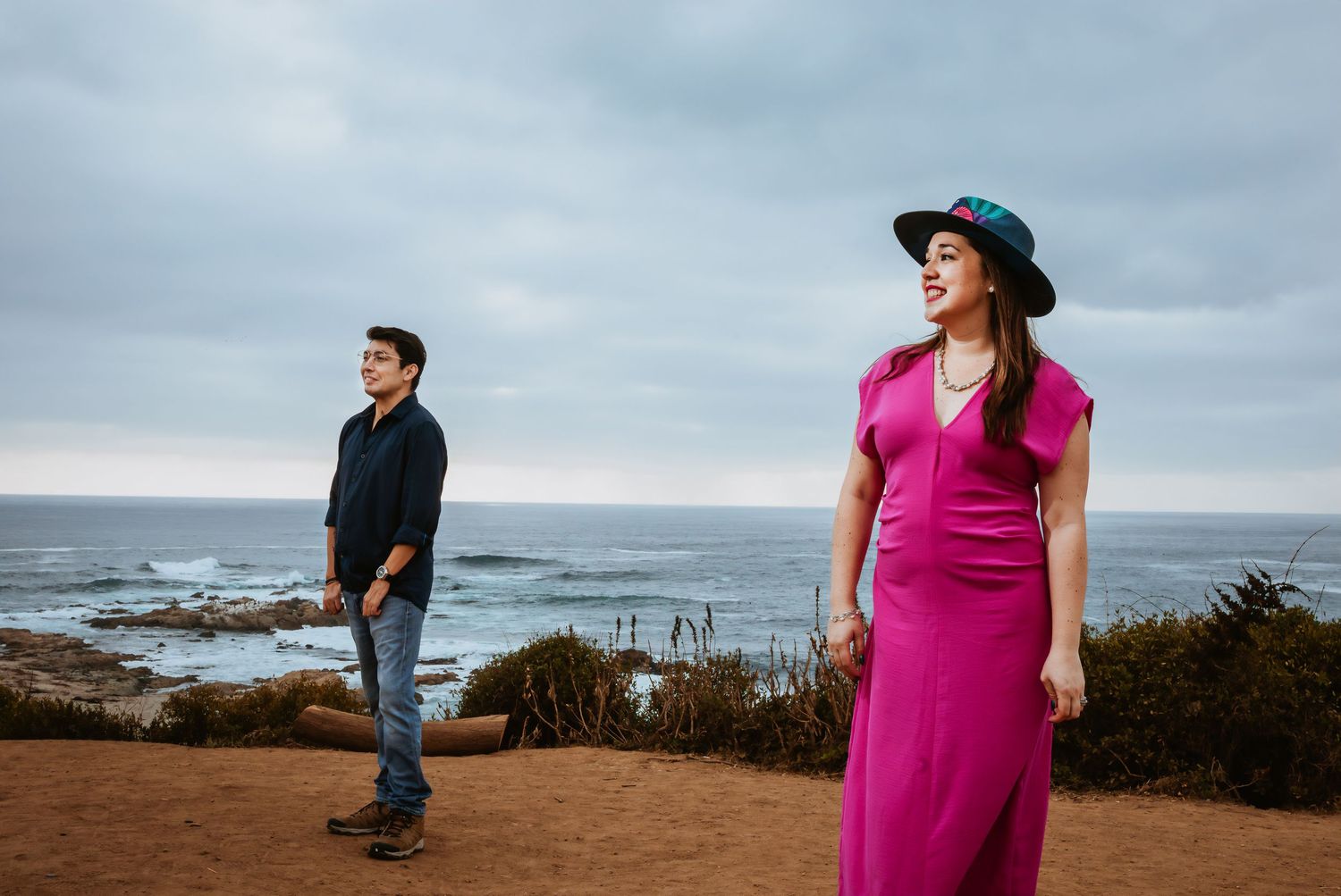 Couple posing on coastal cliffside, woman in pink dress and teal hat, man in navy shirt, ocean backdrop.