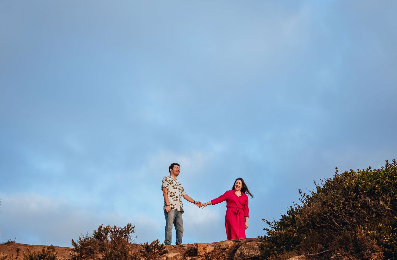 Couple holding hands on a rocky hillside under a blue sky, man in floral shirt and woman in pink dress.