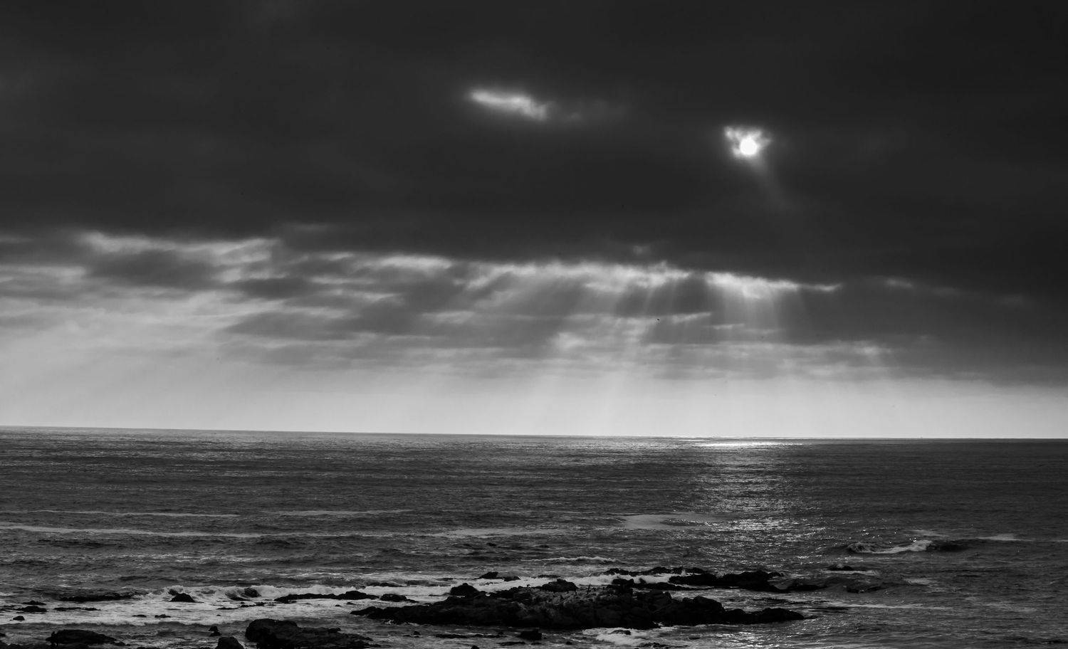 Black and white seascape with dramatic storm clouds and sunrays breaking through over rocky coastline.