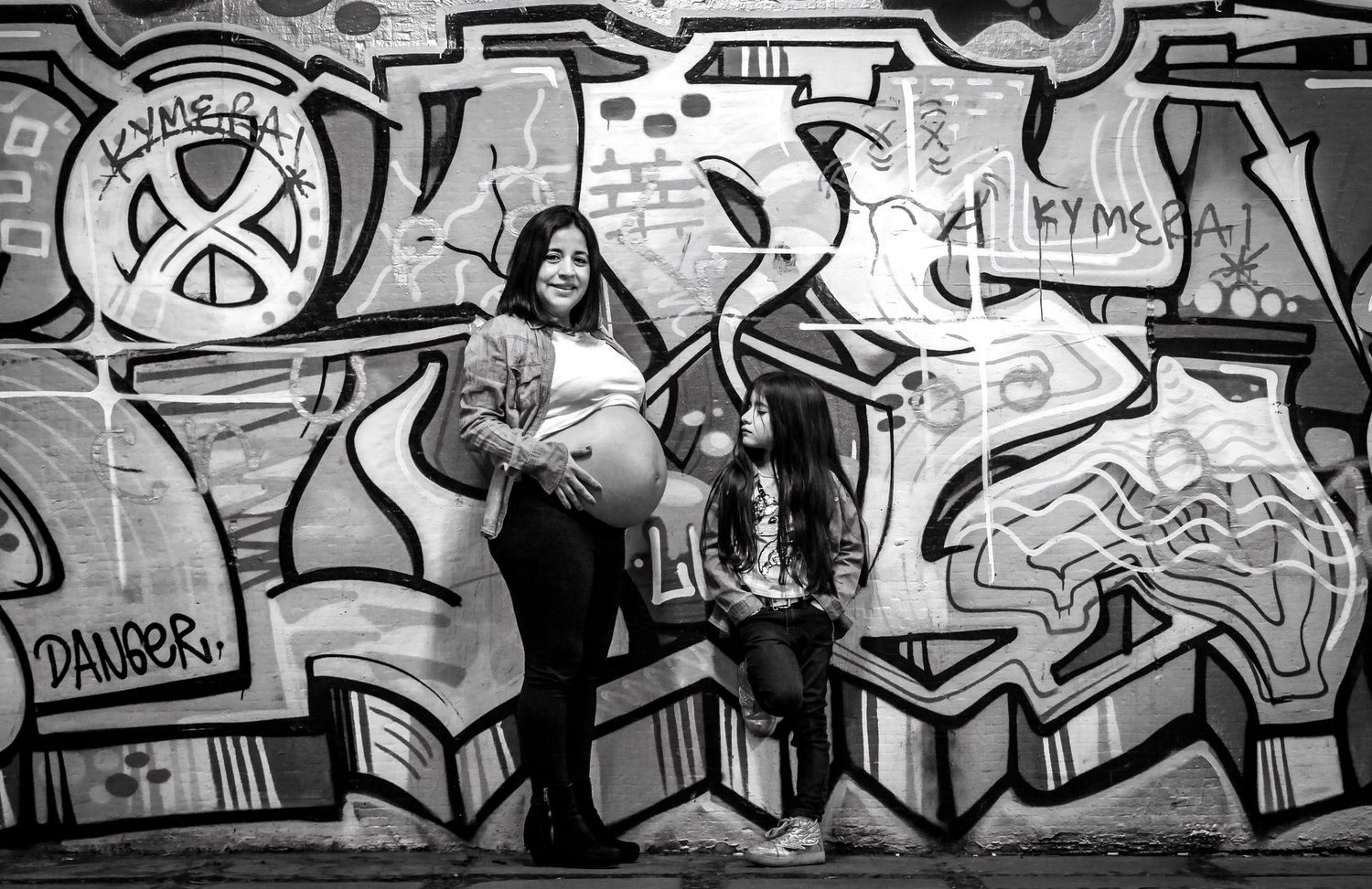 Black and white photo of pregnant woman and child standing before a graffiti-covered wall.