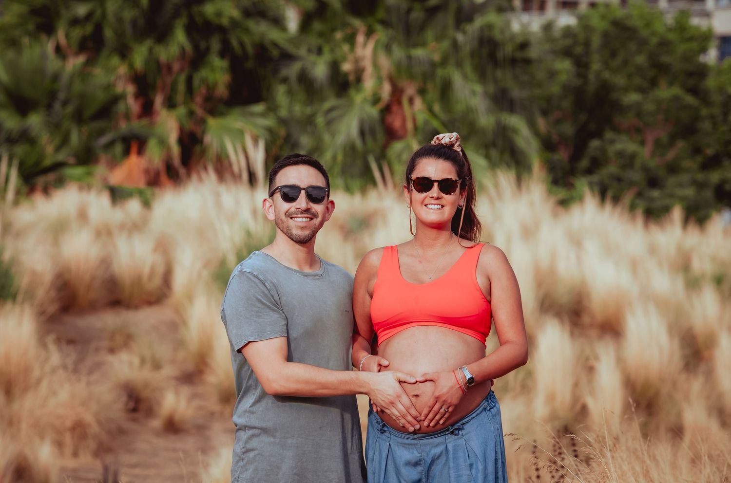 Smiling couple posing together outdoors, man touching pregnant woman's baby bump amid golden grass.