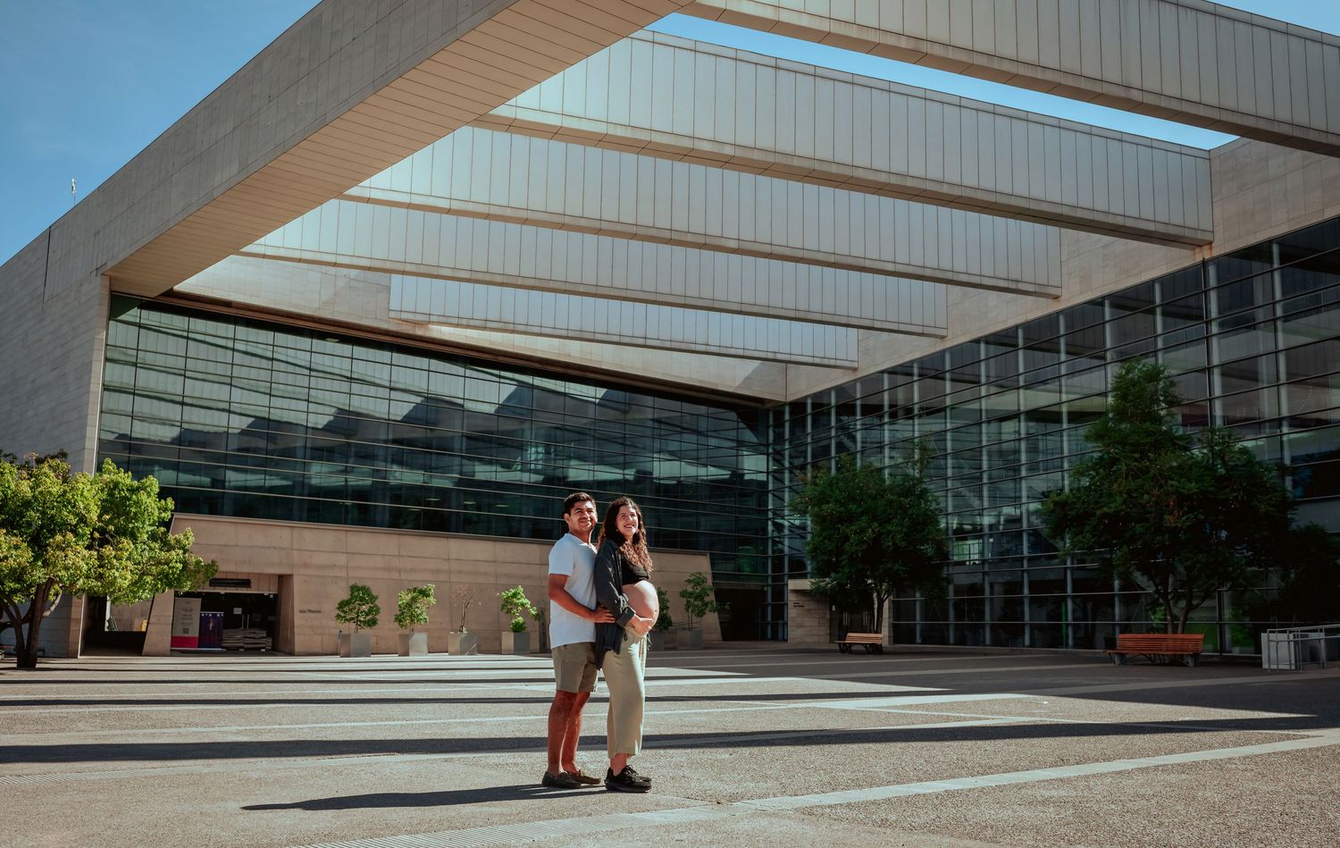 Couple embracing in front of a modern glass building with a large architectural canopy overhead.