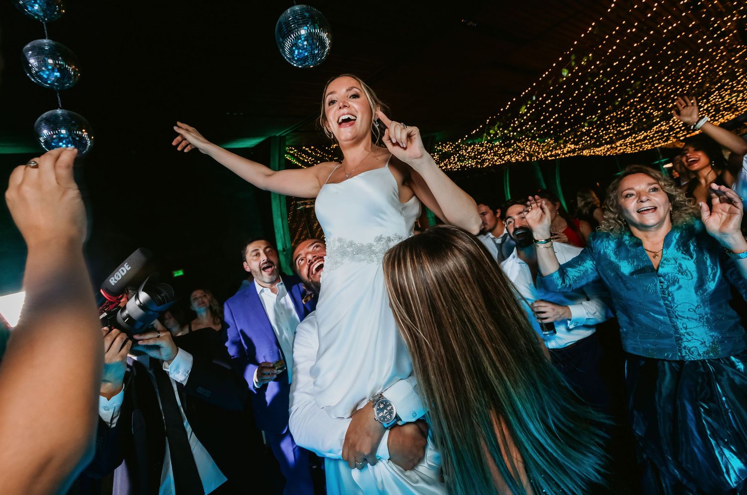 Bride laughing joyfully while being lifted by guests on wedding dance floor.