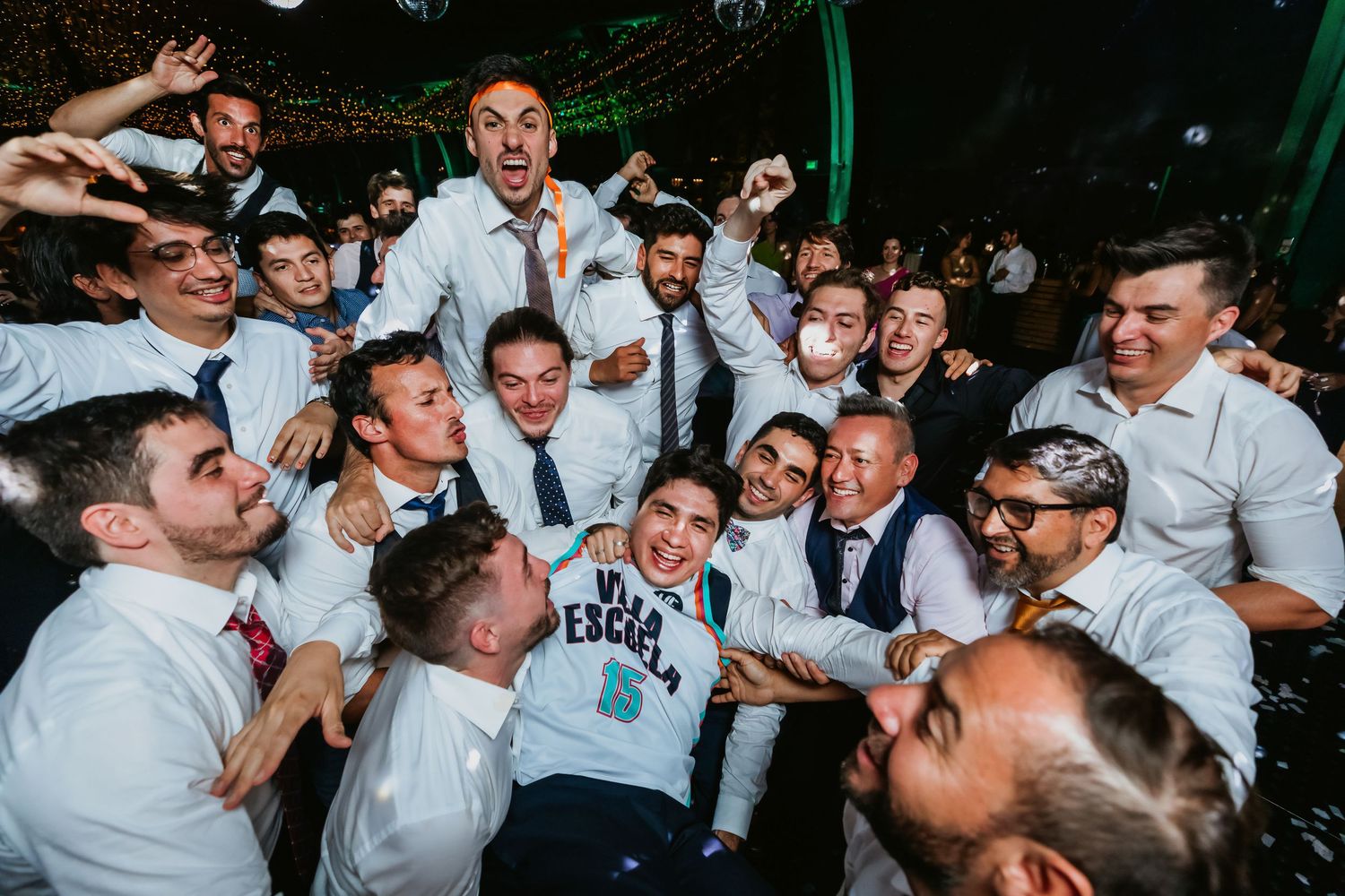 Groom surrounded by celebrating guests in white shirts at energetic wedding party.