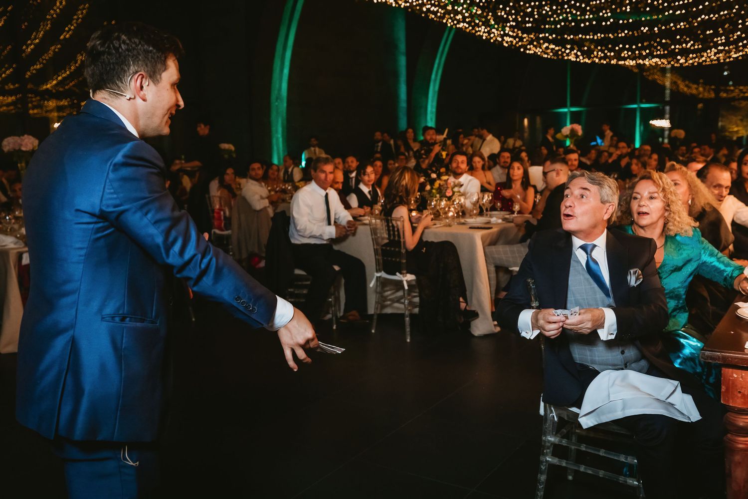 Magician in blue suit performing a close-up magic trick for seated guests at a wedding reception.