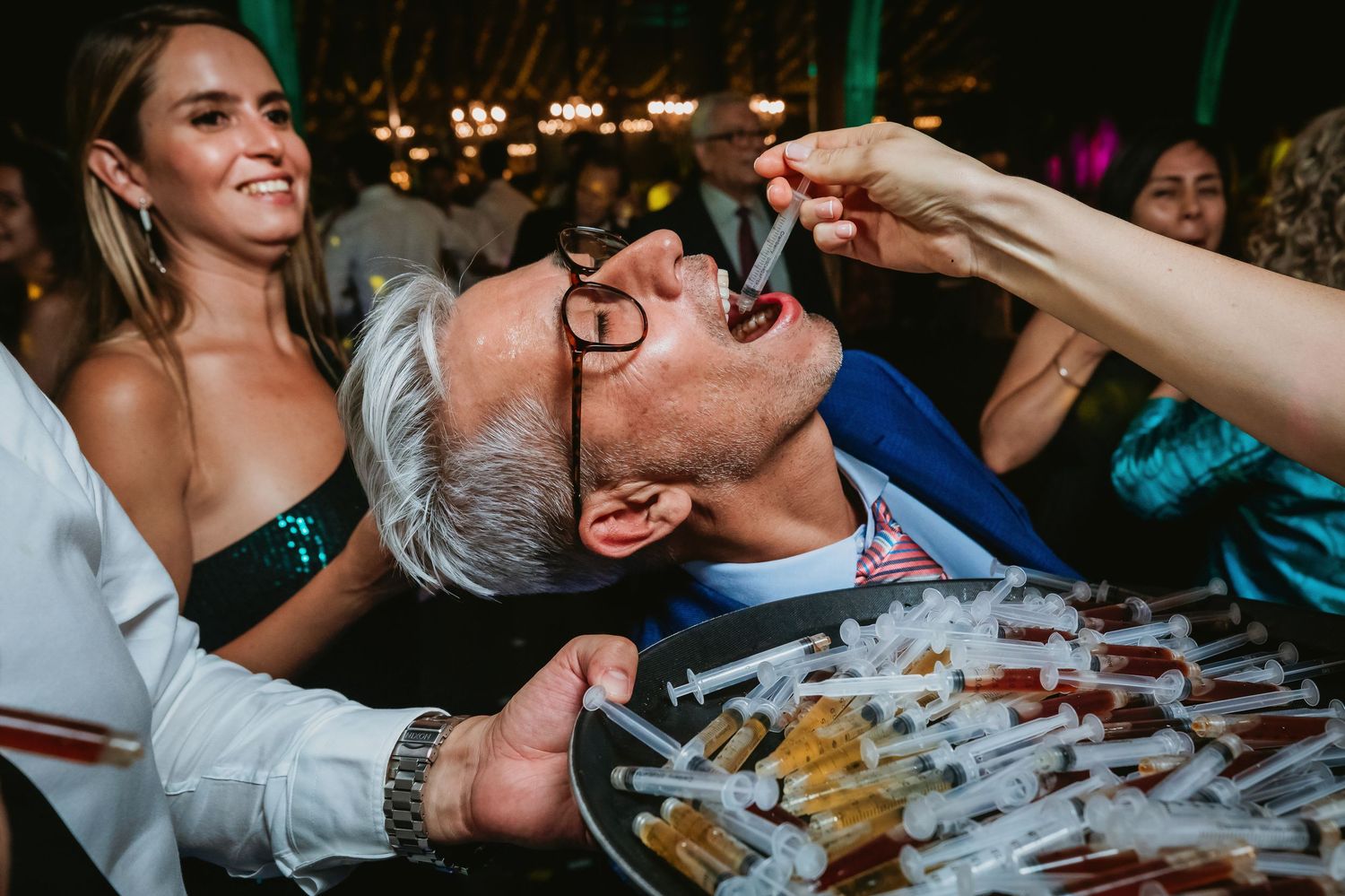 Older man tilts head back to receive a syringe shot from a guest at a wedding.
