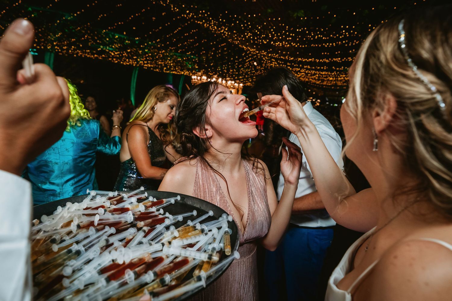 Wedding guest opens mouth as another feeds her a treat from a syringe shot tray.