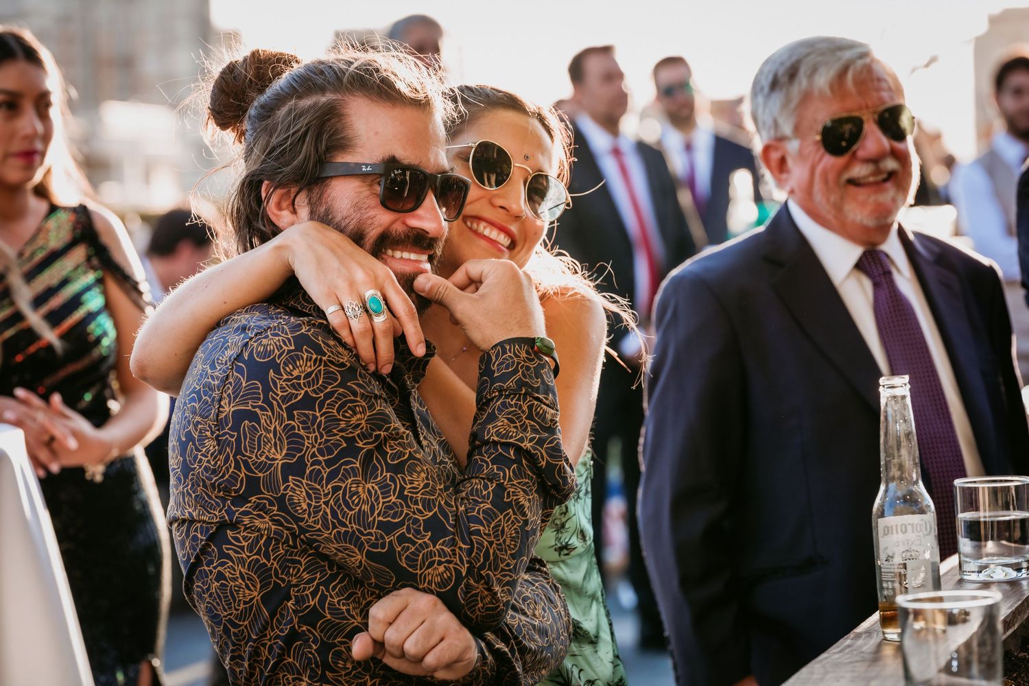Couple in sunglasses embracing and laughing at an outdoor wedding reception.