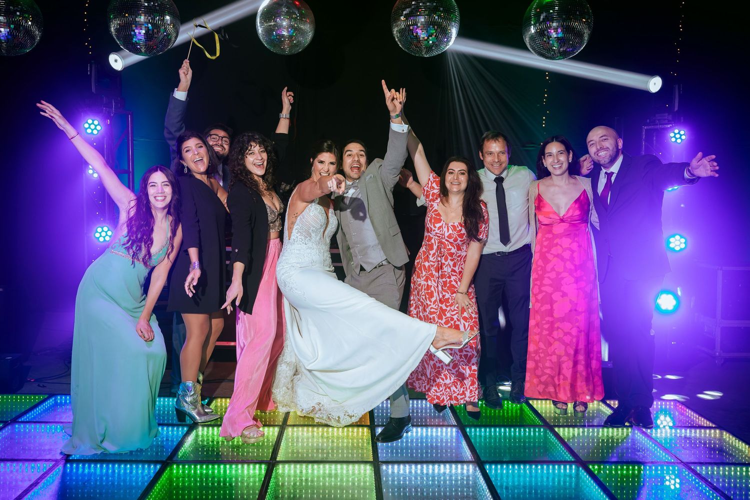 Wedding party celebrating on a colorful LED dance floor under disco balls at a vibrant reception venue.