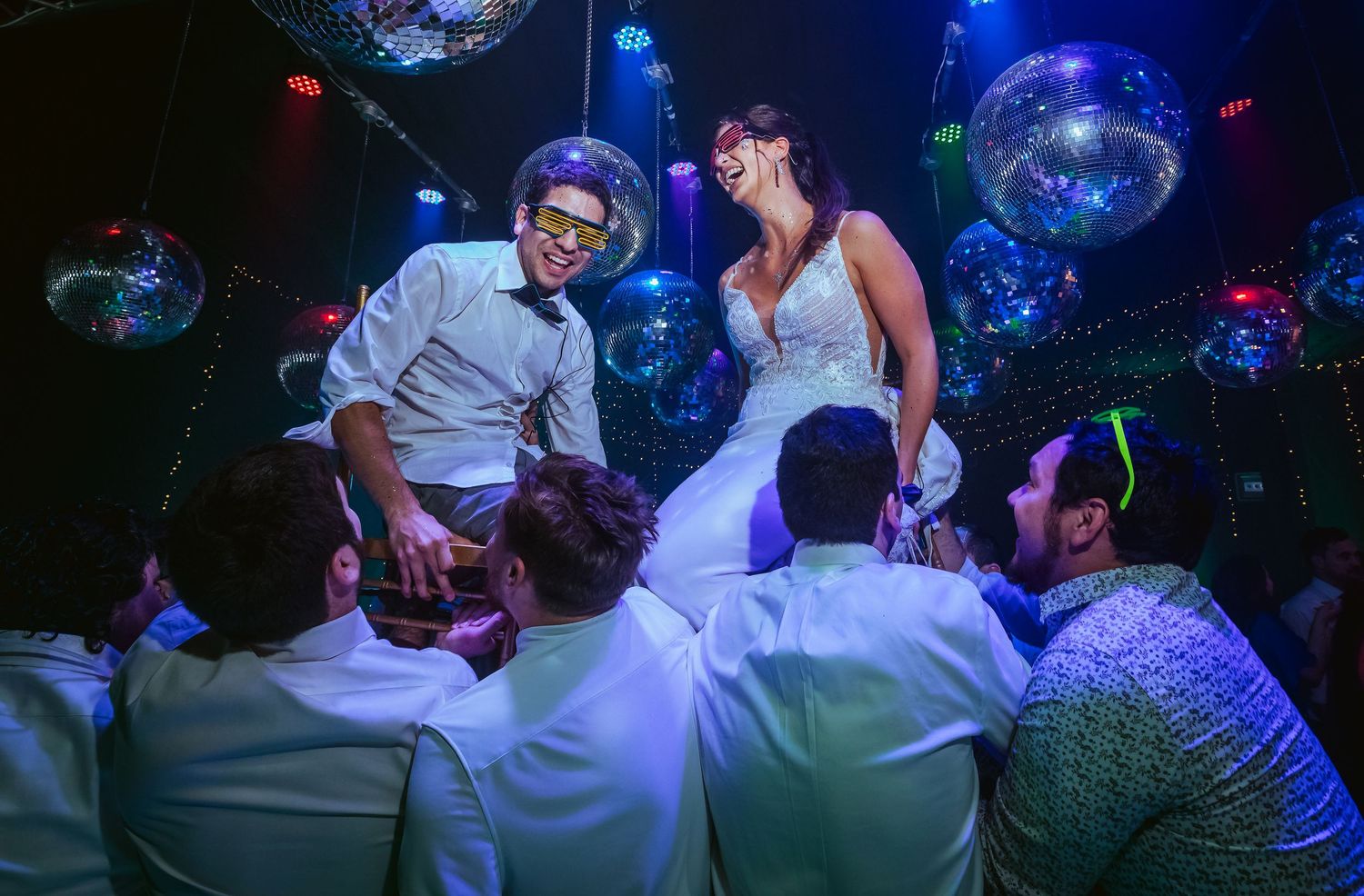 Bride and groom lifted by guests during lively wedding reception with disco balls and colorful lights.