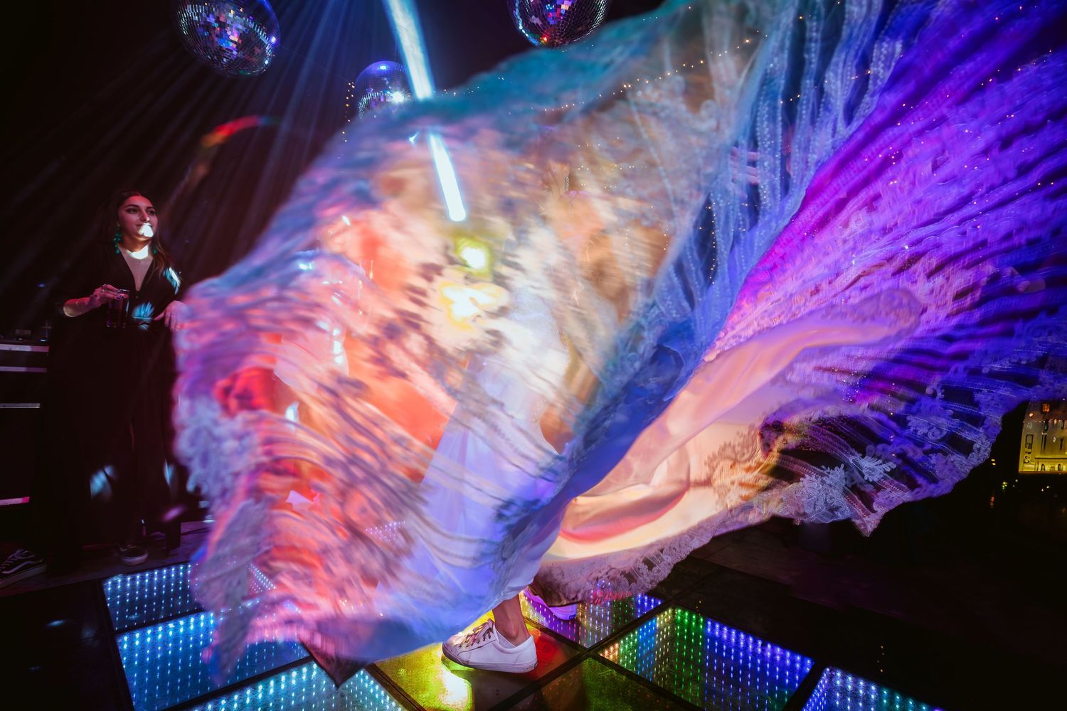 Performer in mask waves giant feathered fans under colorful stage lights on a glittering dance floor.
