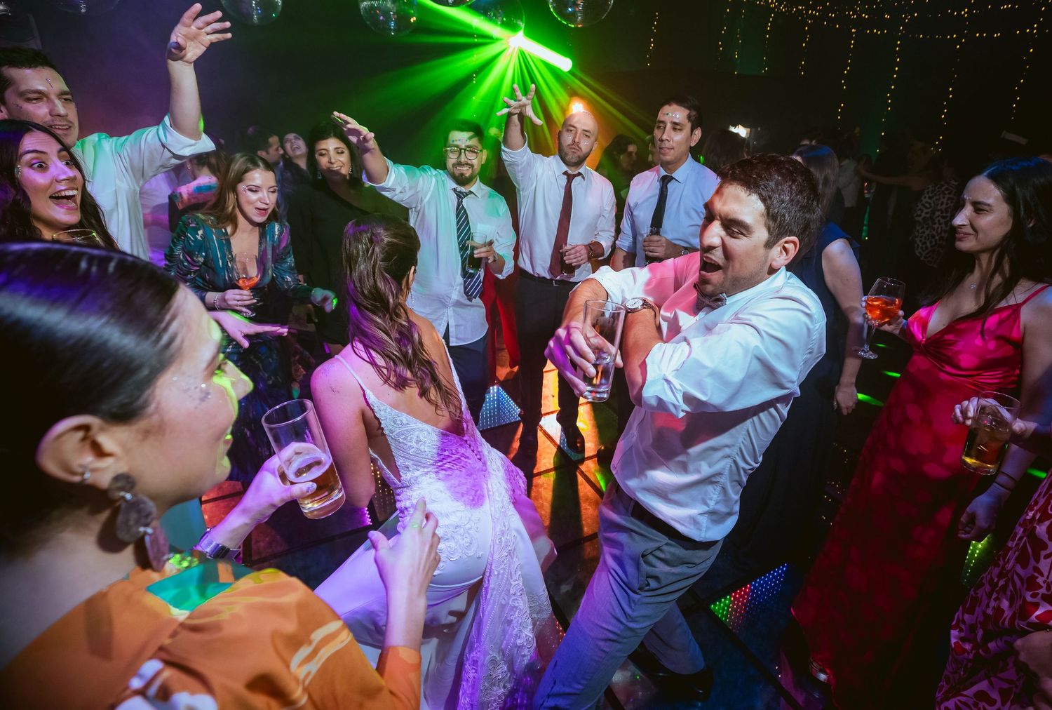 Bride and groom dancing joyfully with guests at a lively wedding reception under colorful lights.