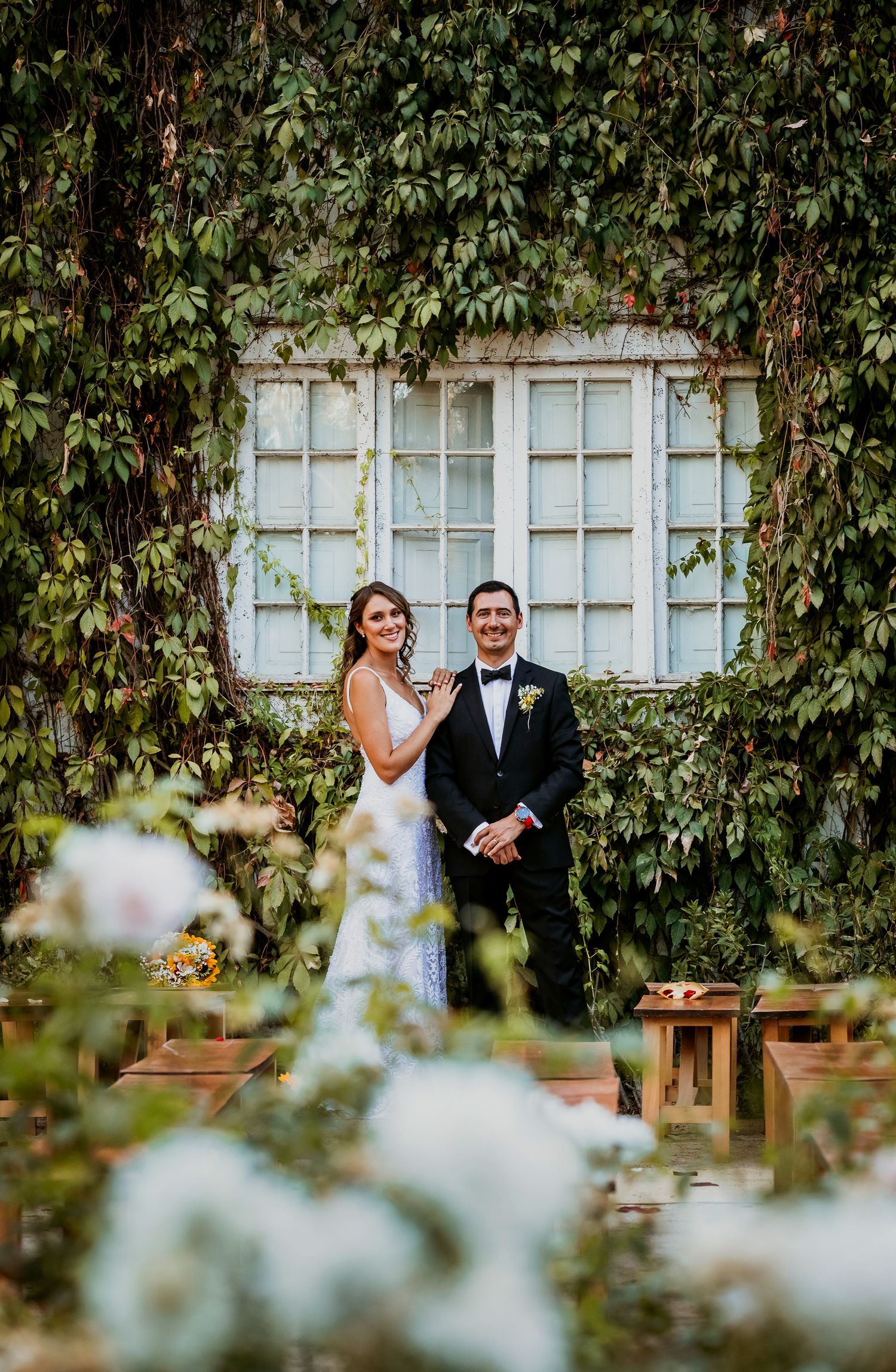 Bride and groom smiling together in an ivy-covered garden courtyard with white floral arrangements in foreground.
