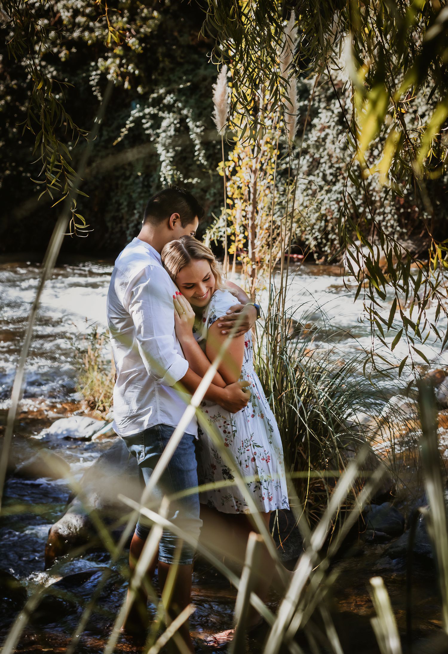 A couple embraces tenderly by a sunlit stream, surrounded by lush greenery and tall grasses.
