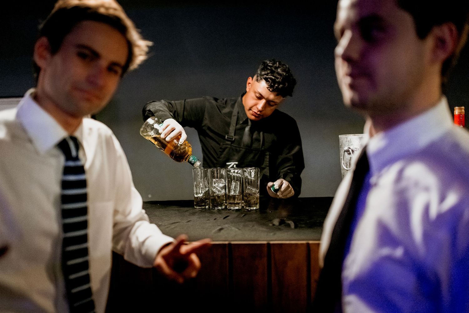 Bartender pouring drinks into glasses while two men in dress shirts and ties watch at a dimly lit bar.