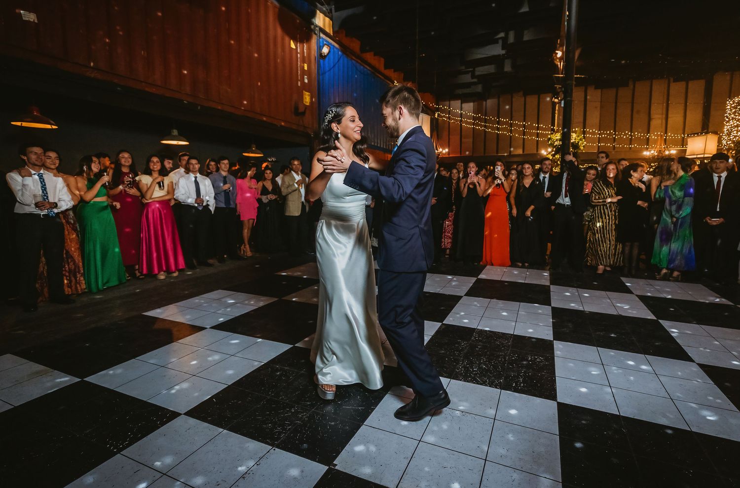 Bride and groom share first dance on checkered floor surrounded by wedding guests in formal attire.